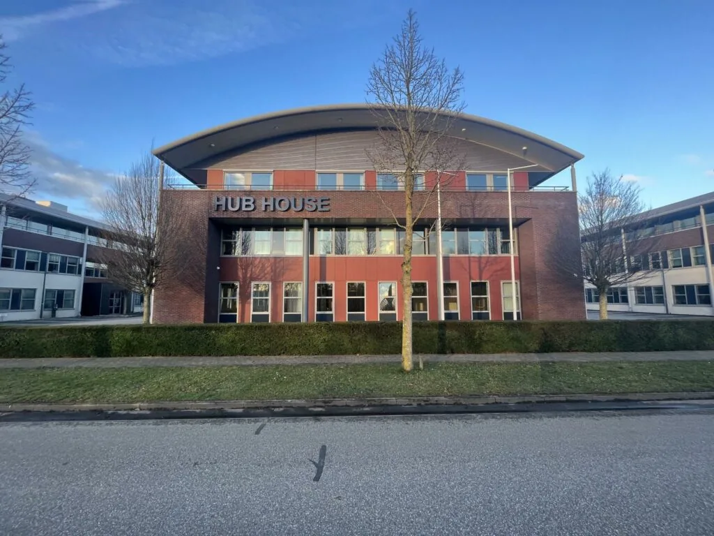 Vooraanzicht van het kantoorgebouw "HUB HOUSE" met rode en bakstenen gevel en kale bomen ervoor.